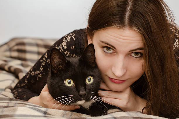Young woman with long brown hair lies on plaid blanket beside black cat with white patch. Both share calm moment, eyes directed at camera, soft indoor light enhancing scene
