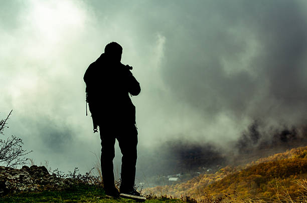 Young man with dark hair stands on grassy hillside, overlooking misty mountains. Dramatic clouds form backdrop, casting shadows on landscape. Scene evokes solitude and introspection