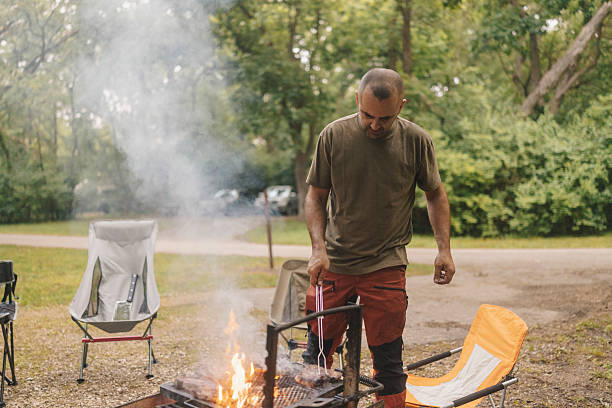 A man of South Asian descent cooks over a fire pit at a rustic campground surrounded by lush greenery, camping chairs, and a serene outdoor setting.