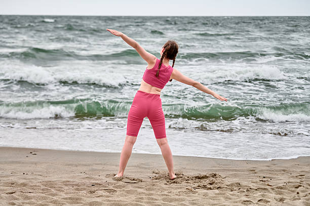 Young woman with brown hair performs yoga on sandy baltic sea beach. Overcast sky, gentle waves, and serene atmosphere set calming tone. Vibrant pink attire contrasts with gray backdrop