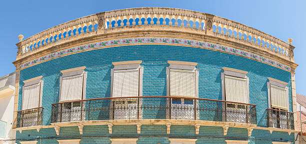 Curved facade of a vintage building covered in blue tiles with closed shutters, floral trim, and wrought iron balconies.