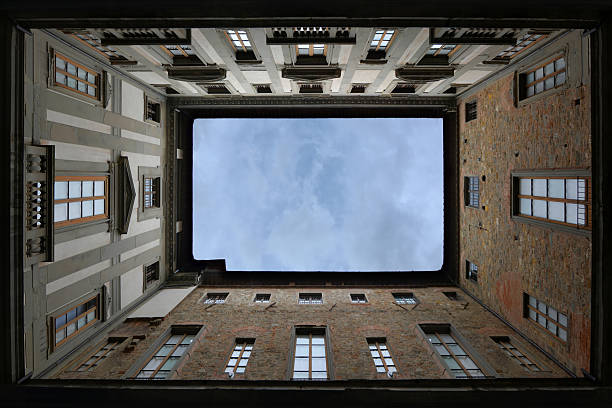 Architectural photograph looking upward from a historic inner courtyard in Florence, Italy. The facades of old stone and stucco buildings frame the rectangular view of the sky above, with dramatic cloudy weather adding depth and mood. The image highlights Renaissance and medieval Italian urban architecture, geometric perspective, and architectural symmetry.
