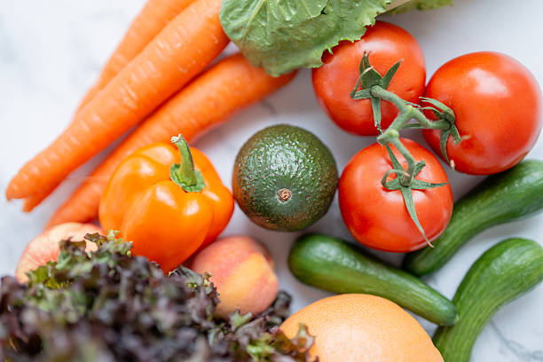 A vibrant assortment of fresh vegetables and fruits including tomatoes, carrots, lettuce, and cucumbers placed against a bright background.
