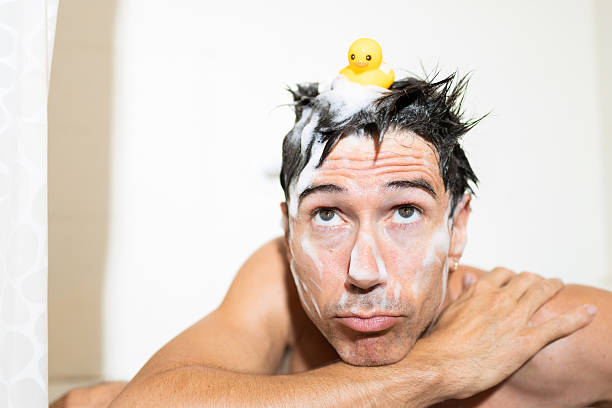 Mid adult man relaxing in a bathtub, soap covering his face, and a yellow rubber duck perched on his head, radiating a sense of boredom while enjoying bath time