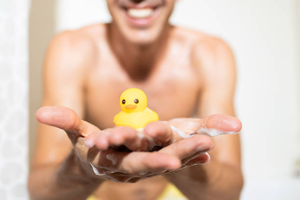 Smiling mid-adult man holding a yellow rubber duck while enjoying a bubble bath, relaxing in a foam-filled tub and embracing a moment of personal care and happiness