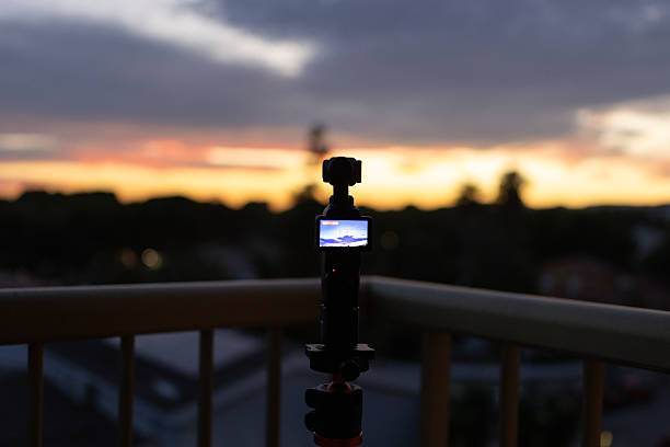 Modern video camera mounted on a sturdy tripod, capturing a stunning timelapse of a vibrant sunset sky from a cozy balcony, showcasing the beauty of evening colors blending in the horizon