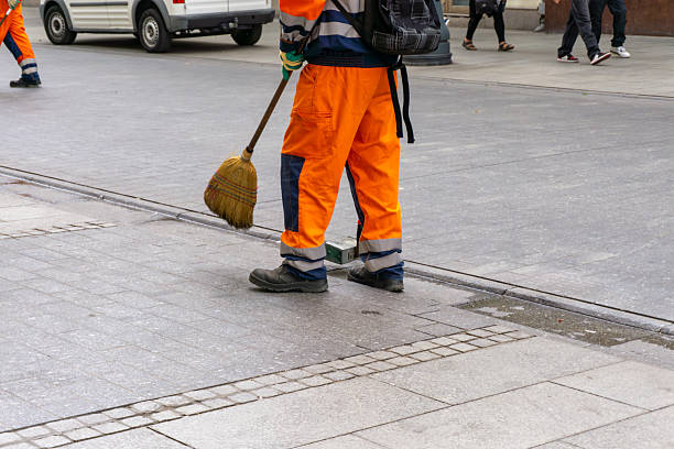 Municipal worker in orange reflective uniform sweeping sidewalk with broom, car and pedestrians visible in the background