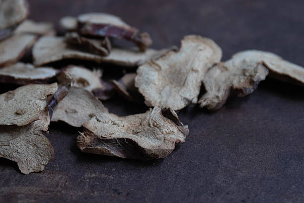 A black bowl filled with Galangal slices, with additional pieces scattered on a dark textured surface. The earthy tones and organic shapes highlight the natural, rustic character of the dried herbs, often used for traditional remedies, cooking, or decorative arrangements