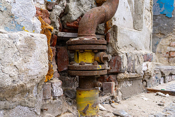 Old gas connection with yellow pipe and rusty valve, installed in damaged brick wall of deteriorating building structure.