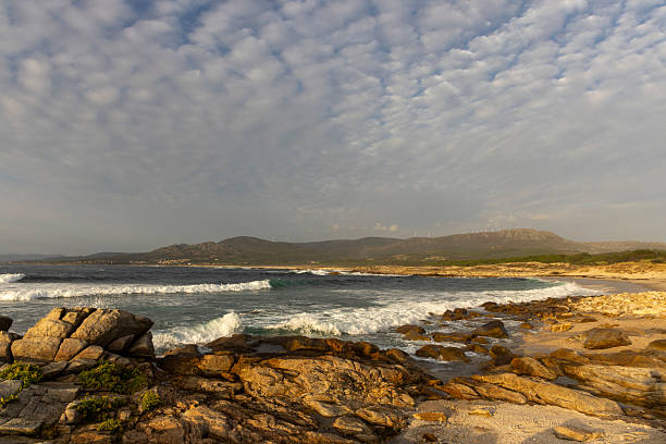 A rocky shoreline with a cloudy sky in the background. The sky is filled with clouds, and the ocean is rough with waves crashing against the rocks. The scene is serene and peaceful, with the ocean