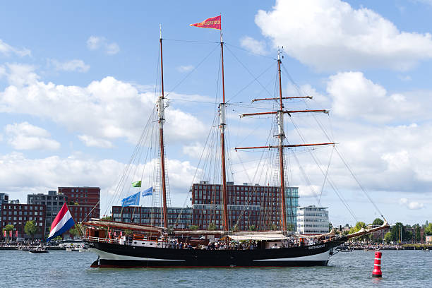 The majestic three-masted tall ship OOSTERSHELDE sails near Amsterdam's city waterfront, capturing the contrast between historic seafaring heritage and modern city life, with thousands of people in audience. During SAIL Amsterdam 2025.