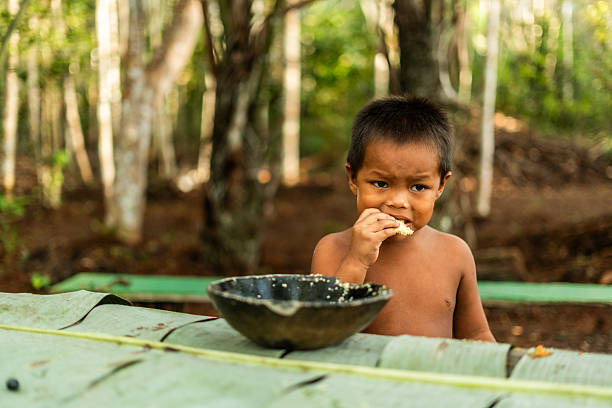 Curripaco child boy community member enjoys traditional meal in handcrafted ceramic utensil under Amazonian vegetation, representing cultural continuity and transmission of ancestral eating habits to new generations in authentic communal context of indigenous life