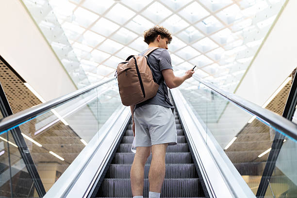 Young Caucasian man with backpack using smartphone while standing on airport escalator. Symbol of mobility, modern travel, youth lifestyle and connection