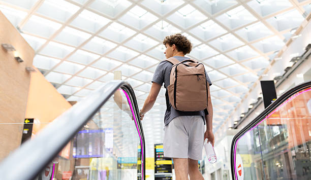 Young Caucasian man with backpack standing on airport escalator, looking back. Concept of journey, modern travel lifestyle, mobility and passenger routine