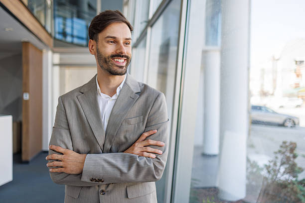 istock Confident businessman smiling and looking out large window in modern office building 2235956684