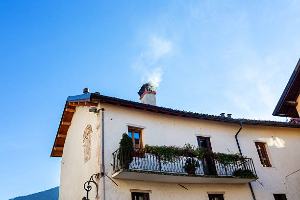 Rustic house features a balcony with plants, smoke rising from chimney, under a bright blue sky, highlighting peaceful countryside architecture and inviting atmosphere