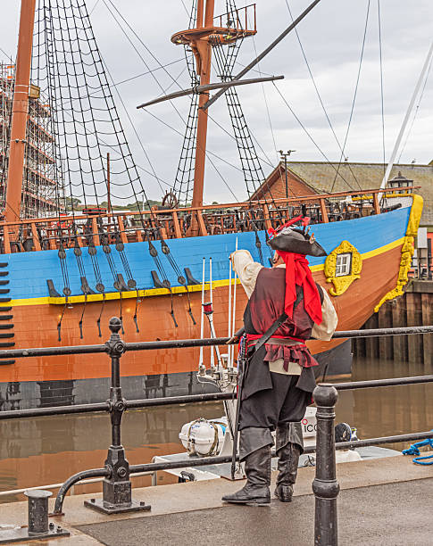 Whitby, UK. September 29, 2025. A man dressed in a pirate costume stands on a quayside. A replica of a historic and colourful tall-ship in the background.