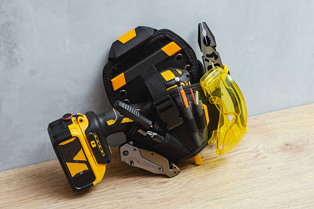 istock Professional construction tool belt with cordless drill, pliers, protective yellow safety glasses, screwdrivers, utility knife, and measuring tools on wooden table. 2235973673