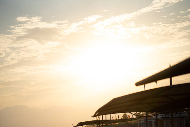 A bright, hazy image with a light yellow sky, scattered clouds, and blurred outlines of beach umbrellas in the foreground and distant mountains.