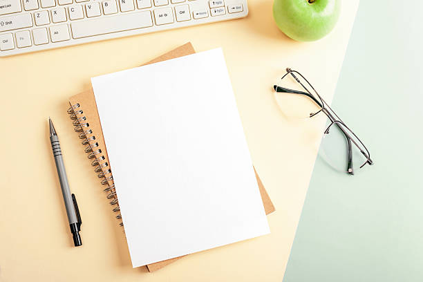 istock White blank paper on spiral notebook with pen, computer keyboard, apple and glasses on yellow and green desk. Top view, flat lay, mockup 2235981432