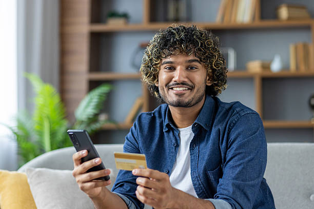 istock Smiling man holding a credit card and smartphone at home 2235983621
