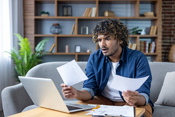 istock Man looking at papers while using a laptop in a modern living room 2235983644