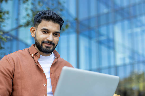 istock A young Indian man with a beard using a laptop outdoors during the day with a smile. 2235986801