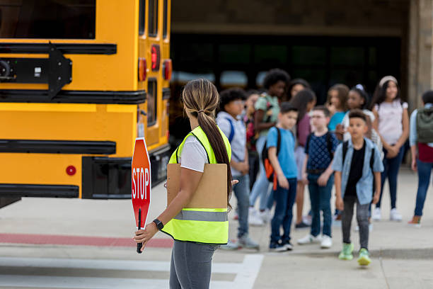 Female crossing guard with stop sign helps children cross near school bus.