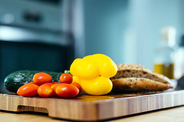 Fresh vegetables including yellow bell pepper, cherry tomatoes, and cucumber arranged on a wooden cutting board with artisanal bread, showcasing healthy cooking ingredients and vibrant colors