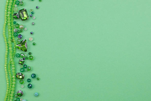 Flat lay top view of beads, crystals, flower-shaped sequins, and rhinestones forming a decorative frame on the left side of a green background. Right side provides ample copy space. Ideal for craft, jewelry, DIY, decoration, advertising, marketing, and banners. High quality photo