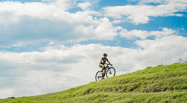 Photograph of a mountain biker climbing a green hill, wearing a helmet and sports glasses, under a blue sky with clouds. Inspiring image that conveys freedom, adventure, and connection with nature.