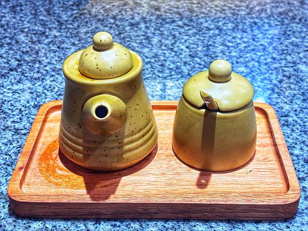 Two earthy yellow ceramic containers with lids, likely a creamer/teapot and a sugar bowl, resting on a wooden rectangular tray against a speckled blue background