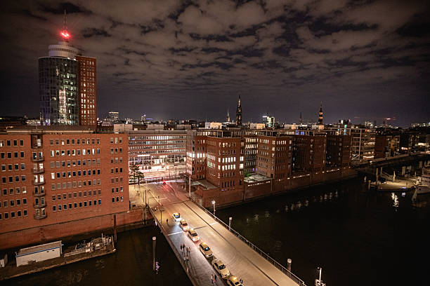 Hamburg, Germany – September 16, 2023: Night view of Hamburg’s HafenCity with illuminated red brick buildings, historic architecture, and the city skyline in the background. The tall tower of St. Nikolai and other church spires rise above the city under a cloudy sky. Reflections of lights shimmer in the canal, showing the mix of heritage and modern city life.