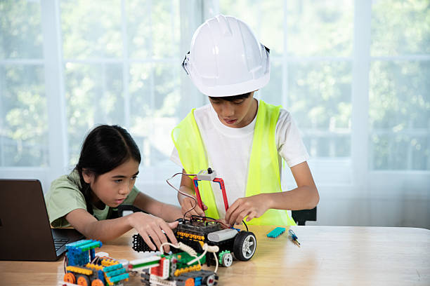 Two Asian children are building a robot car during a STEM class. The boy, wearing a construction helmet and safety vest, works with focus while the girl assists with programming. A great moment of teamwork and hands-on learning in science and technology.