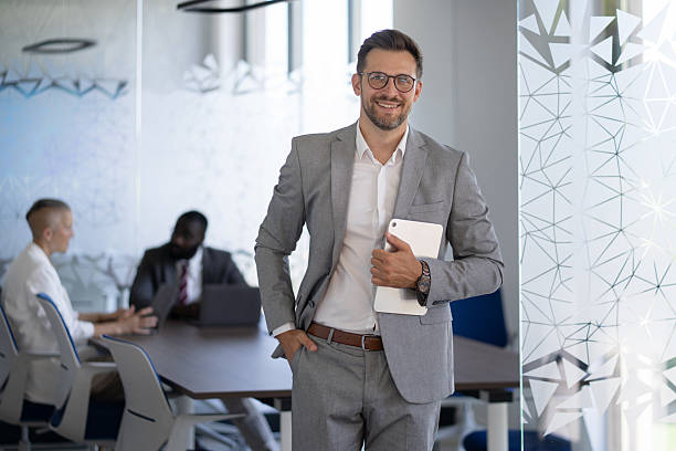 istock Smiling businessman holding digital tablet in modern office with colleagues working in background 2236005540