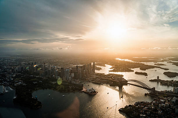 Aerial photograph of Sydney's iconic harbor bridge and opera house with golden sunlight illuminating the city's waterways and urban landscape below.