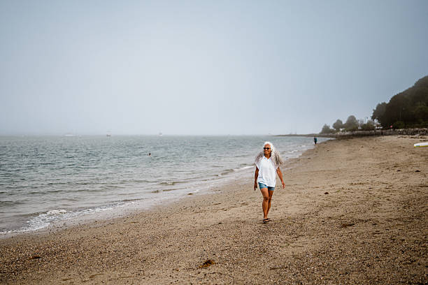 A vibrant multiracial senior woman of Hawaiian and Chinese descent walks leisurely along the peaceful and empty sandy shoreline in Portland, Maine after kayaking with her husband and their friends in Casco Bay.