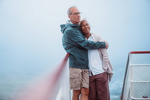 A multiracial senior couple stands on the deck of a ferry in Maine, warmly embracing while enjoying the scenic ride to Peaks Island.