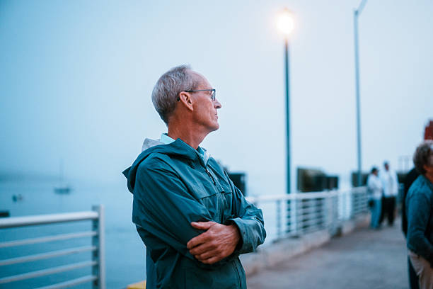 A senior man in a teal jacket stands on a pier in Maine at dusk, waiting to take the ferry back to Portland on a foggy evening.