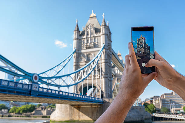 Photographing iconic tower bridge, tourist using smartphone during sunny summer day, capturing london's vibrant urban landscape
