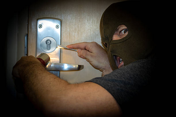 A masked man shining a flashlight on a door that he is trying to open with tools