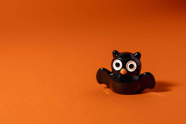 Close-up of a small owl figurine with large eyes casting a dramatic bat-shaped shadow against an orange background. The playful Halloween-inspired setup creates a spooky yet cute atmosphere, ideal for seasonal ads, children's party invitations, autumn holiday designs, greeting cards, and creative campaigns.