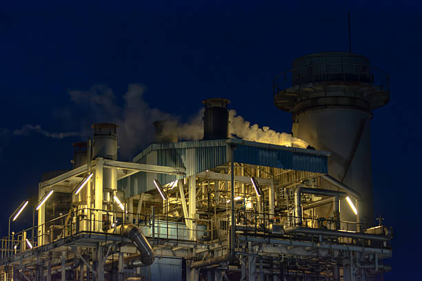 Stock image of a power plant at night, featuring illuminated buildings, pipelines, and industrial structures under artificial lighting. The photo highlights the scale and complexity of the facility, with operational details visible across the site. Perfect for illustrating themes related to industry, electricity production, energy infrastructure, and heavy manufacturing. This image provides a strong visual representation of modern power generation and industrial operations, suitable for use in business, editorial, and environmental content.