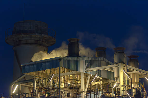 Stock image of a power plant at night, featuring illuminated buildings, pipelines, and industrial structures under artificial lighting. The photo highlights the scale and complexity of the facility, with operational details visible across the site. Perfect for illustrating themes related to industry, electricity production, energy infrastructure, and heavy manufacturing. This image provides a strong visual representation of modern power generation and industrial operations, suitable for use in business, editorial, and environmental content.