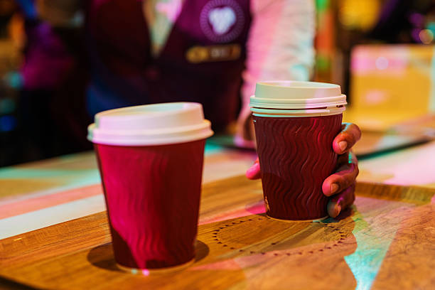 A close-up shot captures a person's hands placing two dark red, textured coffee cups with white lids onto a wooden tray. The action conveys a moment of service at a cafe.