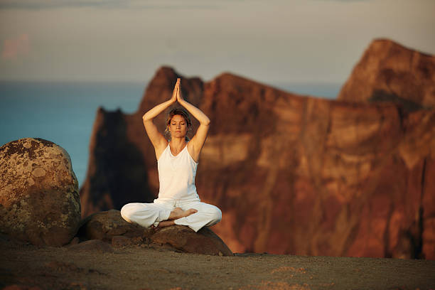 Relaxed athletic woman exercising Pilates with her arms raised during summer day on a cliff above the sea. Copy space.