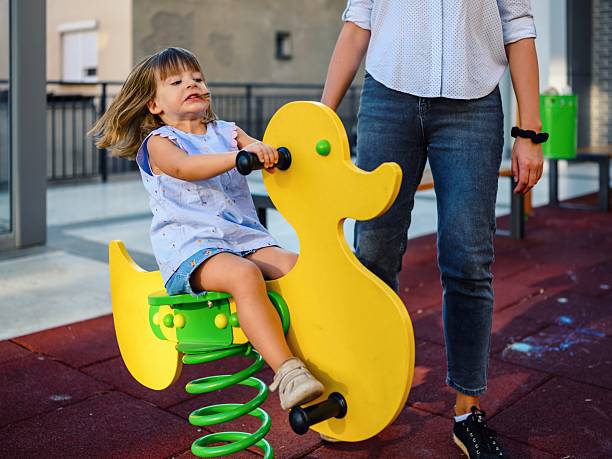 A cute little girl with her mom is using a swing on the kindergarten playground