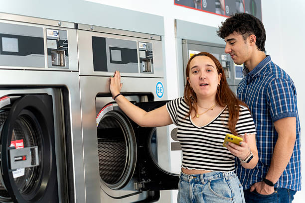 Young woman explaining how to use a washing machine to a young man holding a smartphone in a self-service laundry