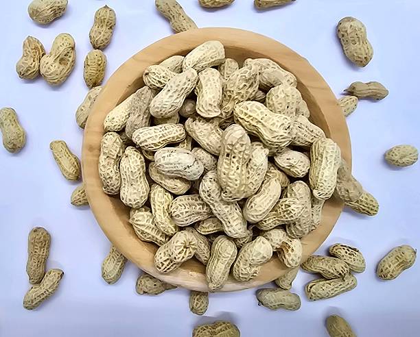 Top view of raw peanuts in shells inside a wooden bowl, with more peanuts scattered on a white background — perfect for food, nutrition, and agriculture themes.