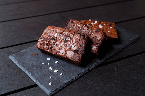 An eye-level shot of two rich, dark, and decadent chocolate brownies with a crackled top. The pastries are elegantly presented on a rectangular slate board, with a sprinkle of white sugar flakes. The high-contrast composition against a dark wooden table highlights the indulgence of this sweet treat.